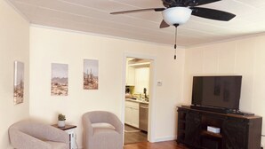 Un soggiorno con ventola a soffitto, una TV su un mobile in legno, due poltrone, un tavolino con una pianta e una porta che dà su una cucina.