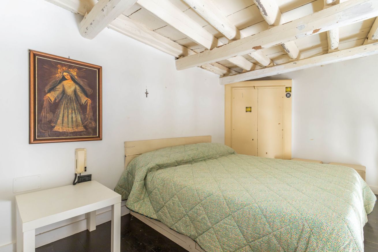 A single bed with a green quilt, a small bedside table, a wall-mounted cross, and a door with a number plate.