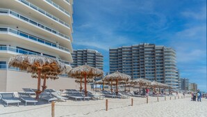 A beach with sun loungers and thatched umbrellas, surrounded by high-rise buildings. - Las Palomas Phase II Cabrillo 1506 Exceptional Oceanview 2BR 2BA Unit (Puerto Peñasco)