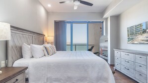 Une chambre d’hôtel dotée d’un grand lit, d’un ventilateur de plafond, d’une table de chevet, d’une commode et d’une grande fenêtre donnant sur l’océan.