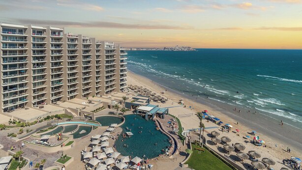 Un hôtel de grande hauteur doté de nombreux balcons, d’un espace piscine donnant sur la plage et d’une plage de sable avec des parasols.