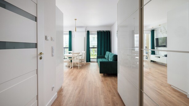 A modern hotel room with a wooden floor, a green sofa, a dining area with a table and chairs, and a TV mounted on the wall. - Bemowo Narwik Deluxe Studio (Warszawa)