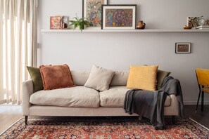 Ein Wohnzimmer mit einem beigen Sofa, einem gemusterten Teppich und Dekorationselementen auf Regalen.