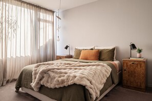 Ein Schlafzimmer mit einem Bett, einem Nachttisch, einem Schrank, einem Fenster mit transparenten Vorhängen und einer Pendelleuchte.