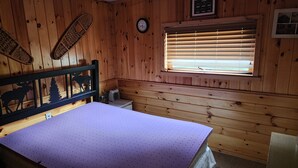 Een houten kamer met een bed, een raam met jaloezieën, een plafondventilator en een wandklok.
