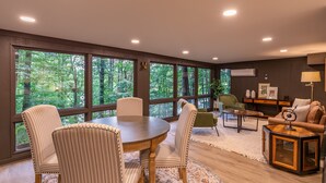 A spacious room with large windows offering a view of a forest, a round dining table with chairs, a sofa, and a small side table.