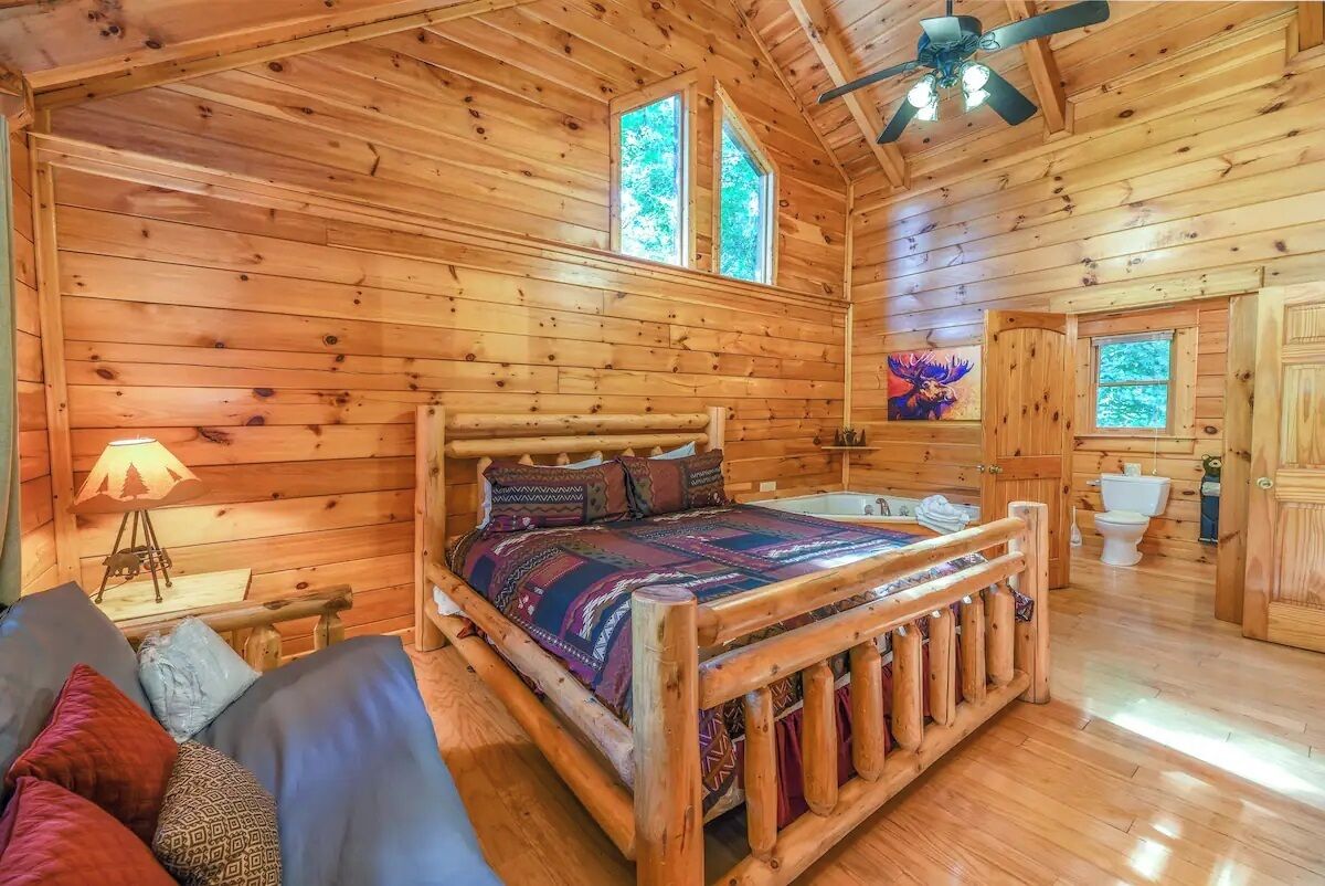 A wooden cabin interior with a bed, a ceiling fan, and a window.