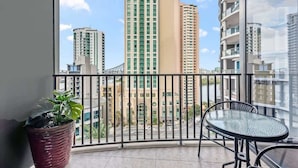 Un balcon donnant sur de hauts immeubles, une plante en pot et une table ronde avec des chaises.