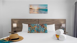 A hotel room with a wooden headboard, a beach sunset painting, a floral pillow, two bedside lamps, a straw hat, a blue towel, and rolled white towels.