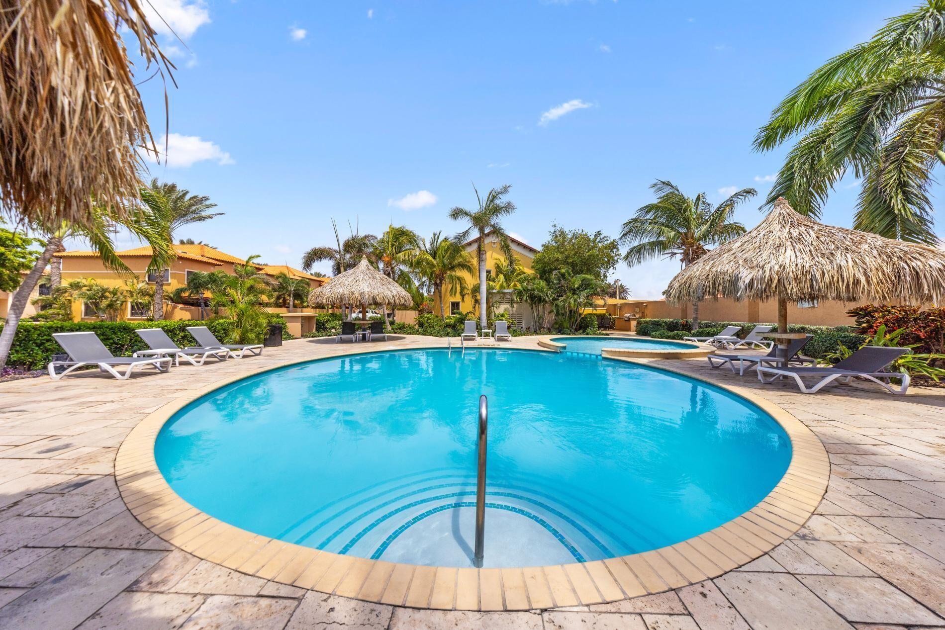 A circular pool with lounge chairs and thatched umbrellas, surrounded by palm trees and a building with a yellow facade.