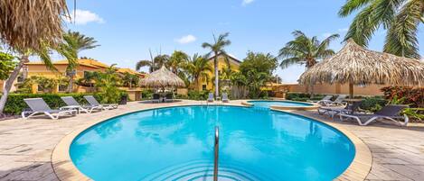 A circular pool with lounge chairs and thatched umbrellas, surrounded by palm trees and a building with a yellow facade.