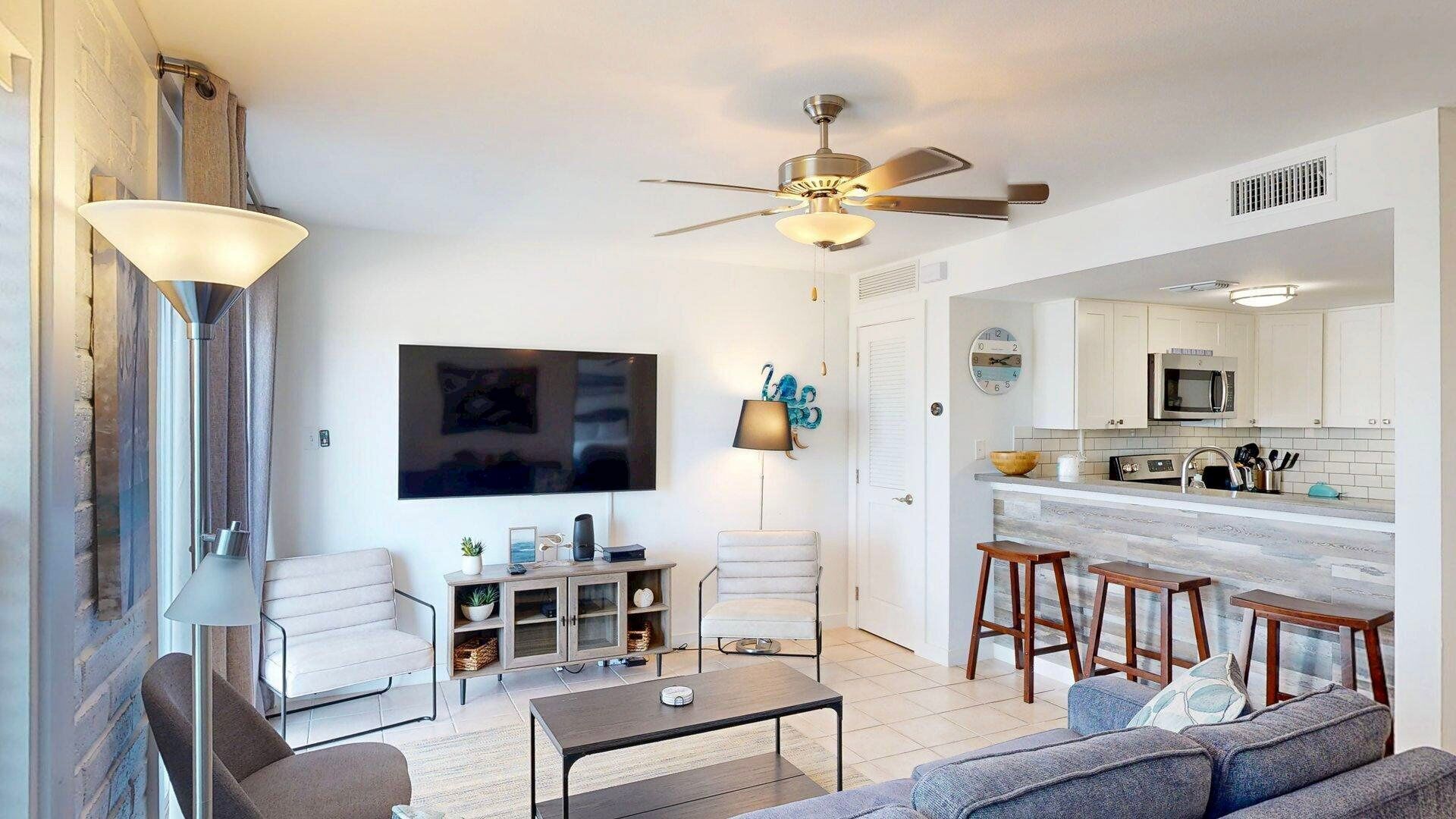Una sala de estar moderna con un sofá, una mesa de centro y un televisor. Hay una zona de cocina con un microondas y taburetes.