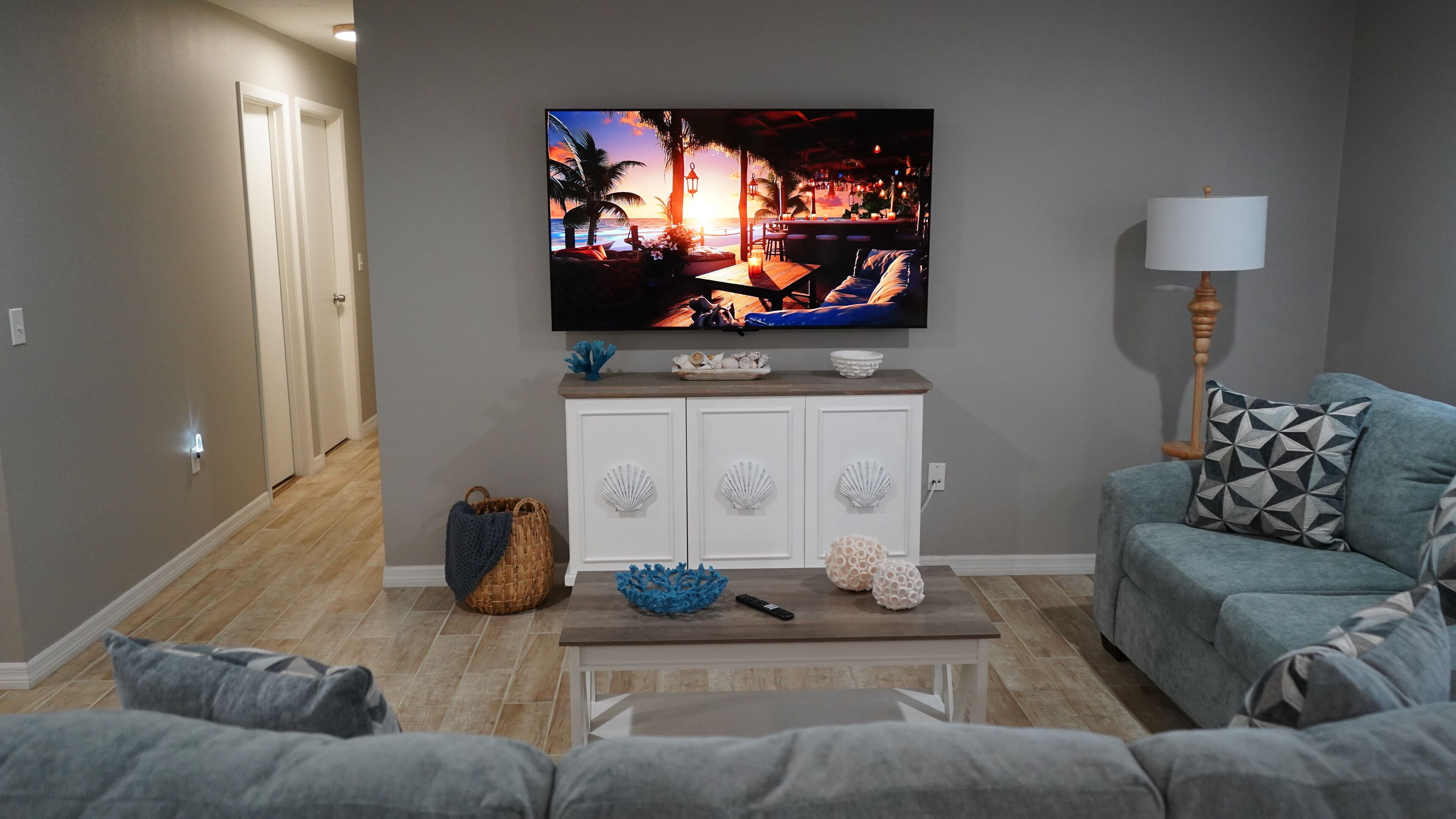 A living room with a flat-screen TV mounted on the wall, a wooden coffee table, a blue decorative bowl, a wicker basket, and a grey sofa with patterned cushions.
