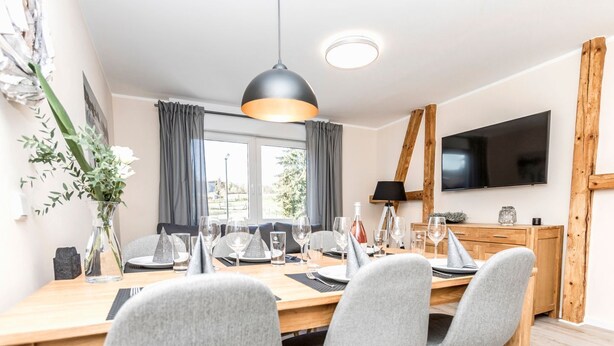 Un moderno comedor con una mesa de madera dispuesta para cuatro, sillas tapizadas en gris, una lámpara colgante y un televisor de pantalla plana montado en la pared.