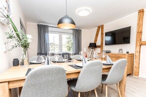 Un moderno comedor con una mesa de madera dispuesta para cuatro, sillas tapizadas en gris, una lámpara colgante y un televisor de pantalla plana montado en la pared.