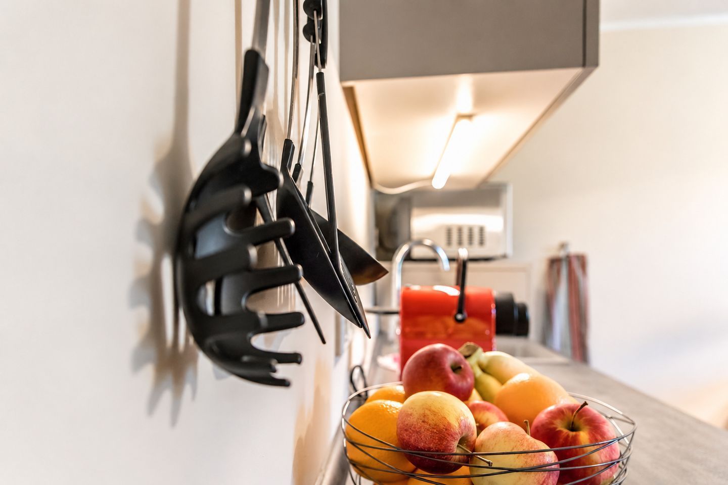 Een keuken met een fruitschaal met appels en sinaasappels, een rode waterkoker en een zwarte grillpan aan de muur.