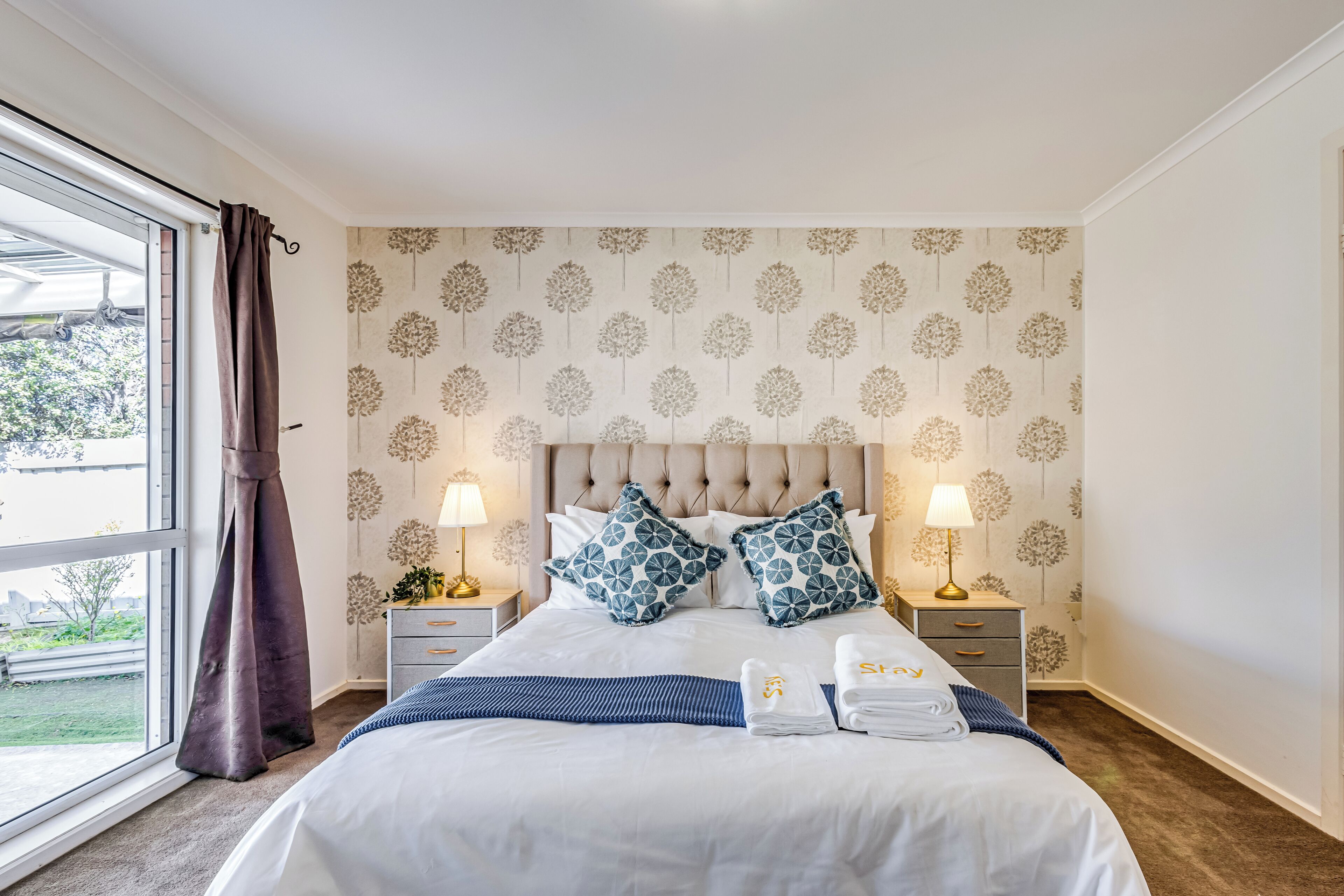 A neatly made bed with a tufted headboard, two bedside tables with lamps, and a view of the outdoors through a large window.
