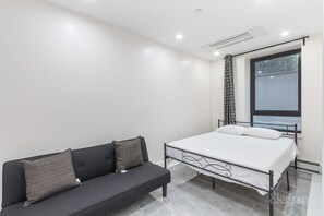 A modern bedroom with a bed, a black sofa with patterned cushions, a large window, and a ceiling light.