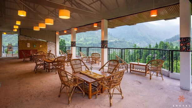 A covered outdoor seating area with wicker furniture and a view of green hills. - Zostel Pulga (Bhuntar)