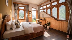 A bedroom with a wooden bed, a desk, and large windows with mountain views. - Zostel Pulga (Bhuntar)