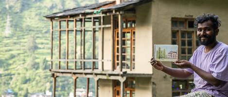 Une personne tient une carte illustrée d’une maison et se tient devant un bâtiment traditionnel doté de grandes fenêtres et d’un toit en pente, le tout sur fond de paysage montagneux.