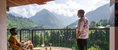 A balcony with a view of mountains, a table with a vase, and two people conversing.