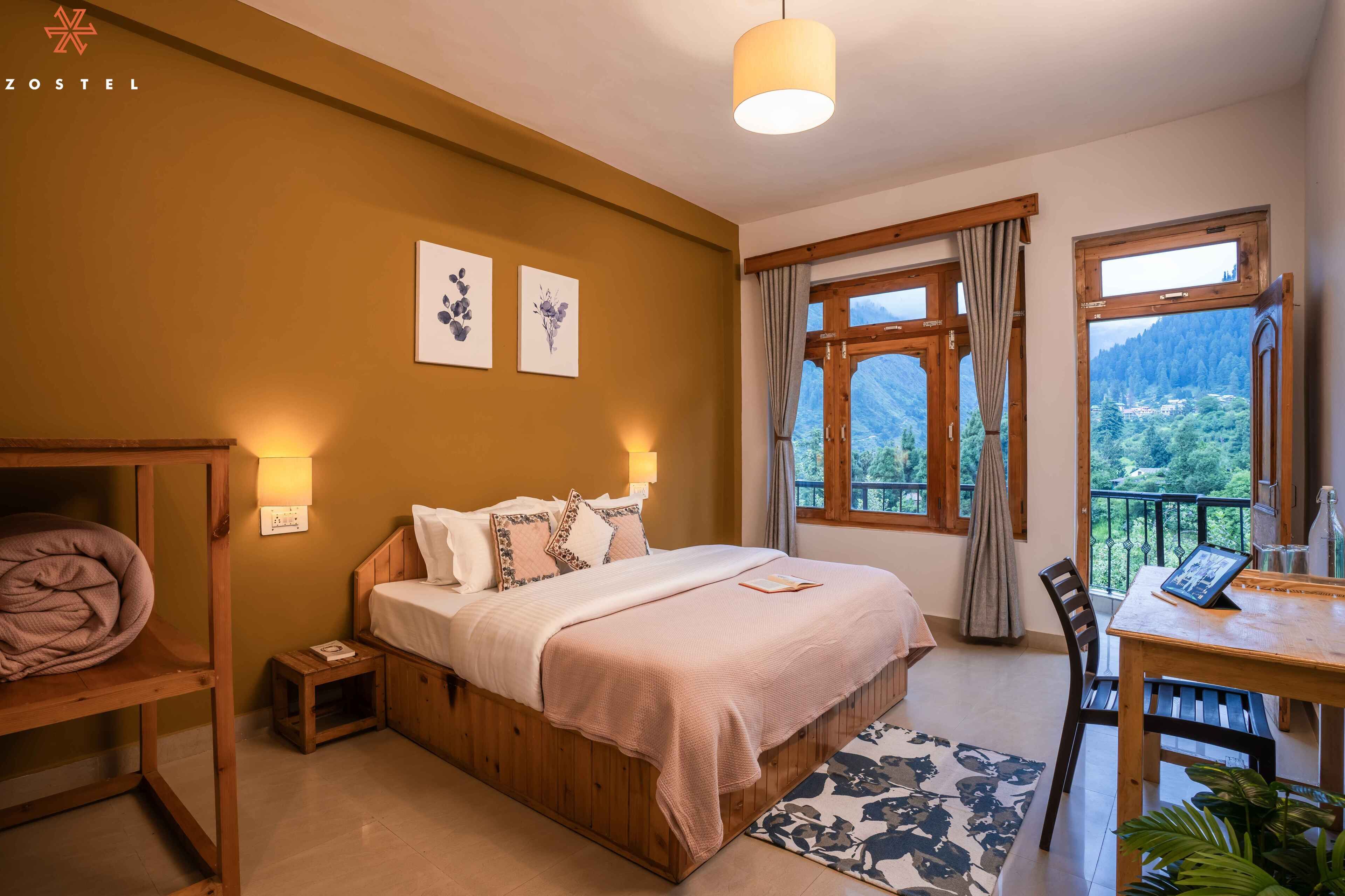 A cozy bedroom with a wooden bed, a small bedside table, a chair, a desk with a laptop, and large windows offering a mountain view.