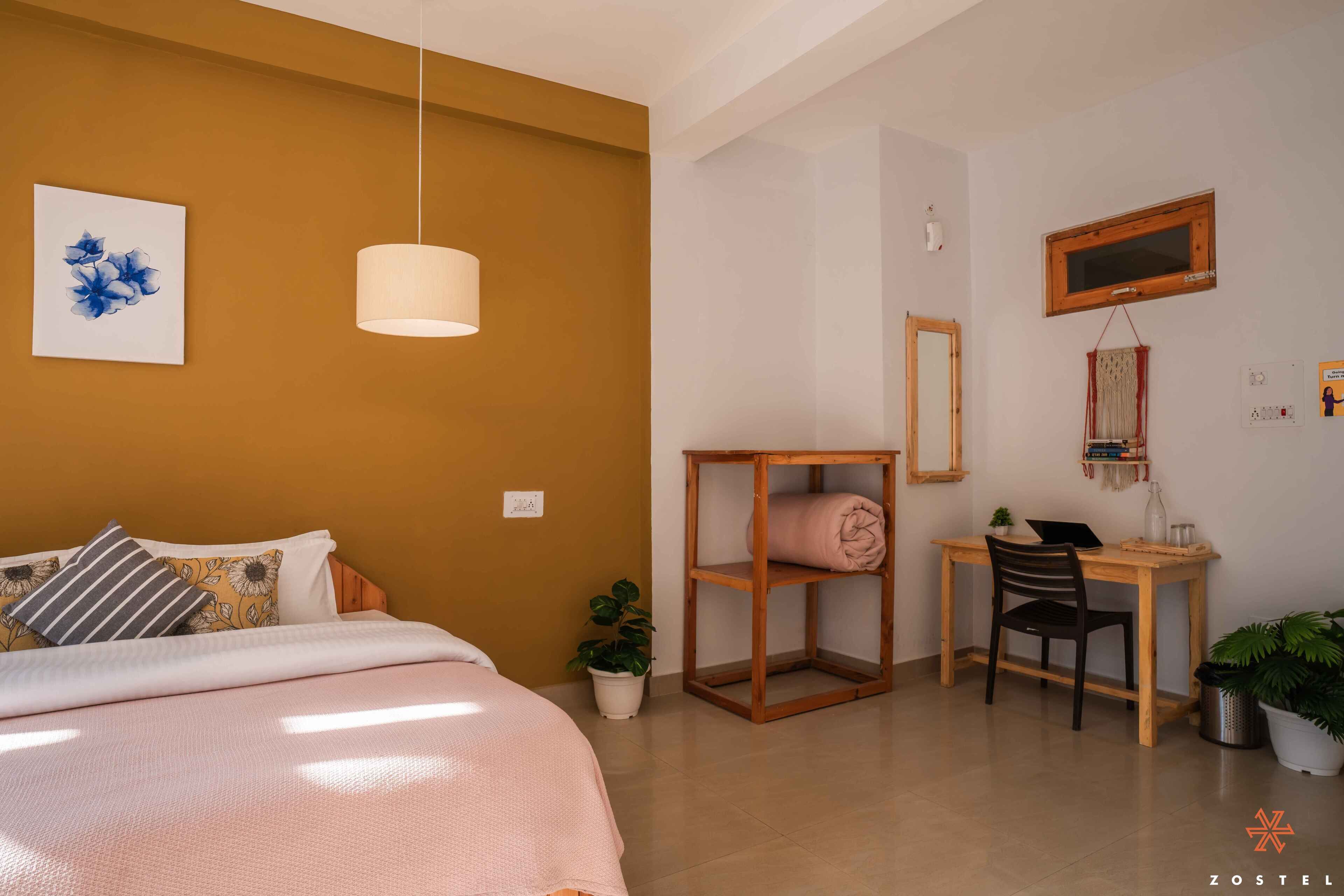 Un dormitorio con cama, mesita de noche y escritorio con silla. Se ve una ventana con marco de madera y un espejo en la pared.