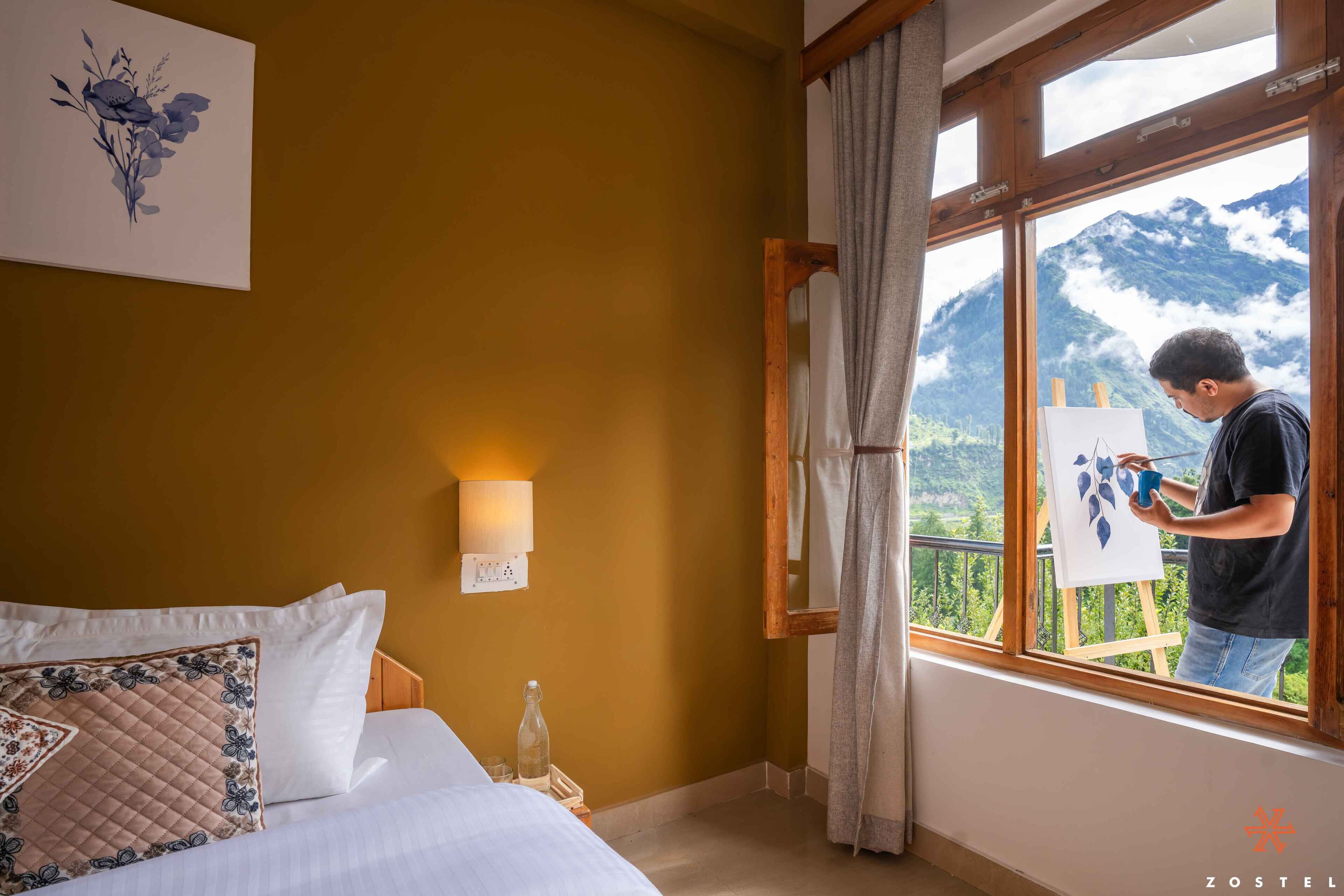 Une chambre d’hôtel avec un lit, un tableau accroché au mur, une fenêtre donnant sur les montagnes et une personne en train de peindre.