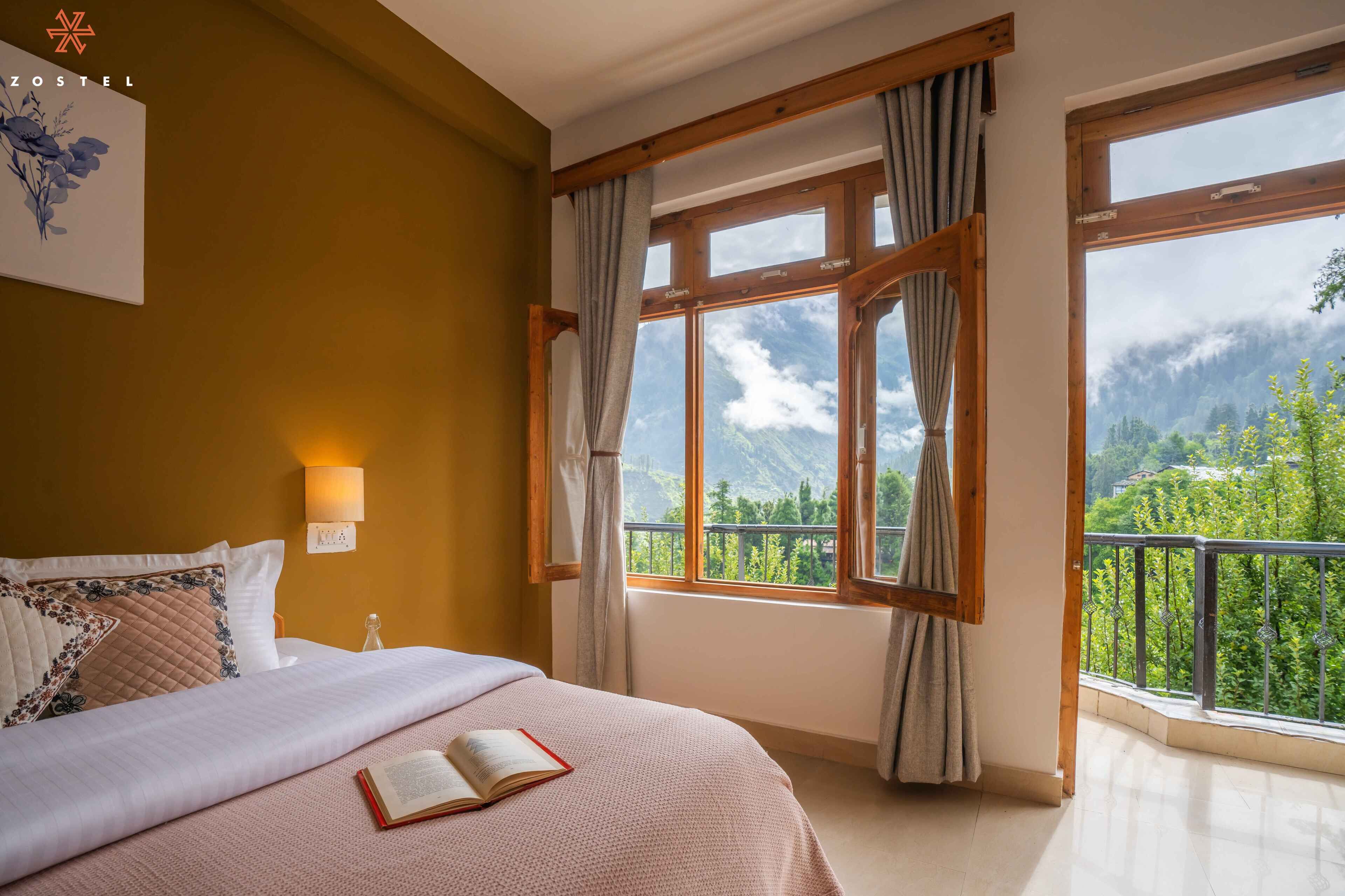 A hotel room with a bed, a window with curtains, a wall-mounted light, and a view of greenery and mountains.