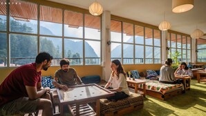 A cozy room with large windows, wooden furniture, and a foosball table. - Zostel Pulga (Bhuntar)
