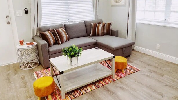 A living room with a gray sectional sofa, a white coffee table, and two yellow ottomans. - Spacious 1-Bedroom in Coral Ridge with King Bed, Sofa Bed & Pool Access (fort lauderdale)