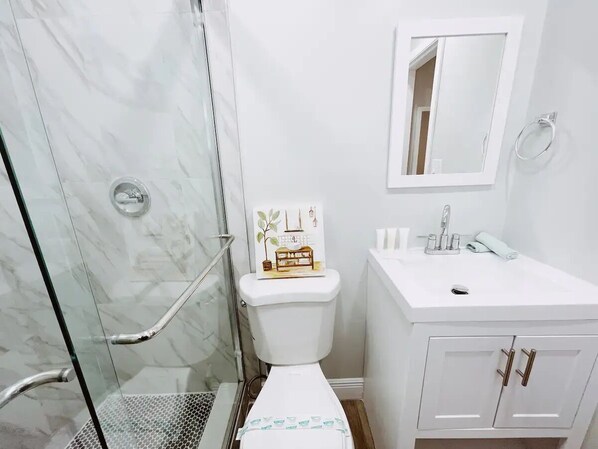 A bathroom with a glass shower enclosure, a toilet with a decorative plaque, a white sink with a mirror above, and a cabinet with two drawers. - Spacious 1-Bedroom in Coral Ridge with King Bed, Sofa Bed & Pool Access (fort lauderdale)