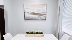 A white table with a wooden planter containing plants, flanked by white chairs, in a room with a white wall and a framed abstract painting above the table. - Spacious 1-Bedroom in Coral Ridge with King Bed, Sofa Bed & Pool Access (fort lauderdale)