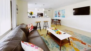 Una sala moderna con un sofá de cuero, una mesa de centro con frutas, y una cocina al fondo.