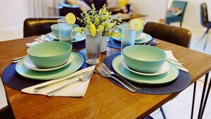 Une table à manger dressée avec des bols, des assiettes et des couverts de couleur turquoise, accompagnée de verres d’eau et d’un vase contenant des feuilles vertes et des tranches de citron.