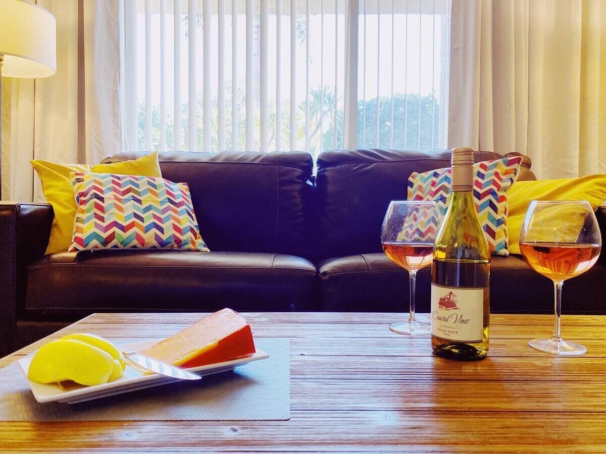 A living room with a dark leather sofa, colorful pillows, a wooden coffee table with a bottle of wine, glasses, and a plate of food.