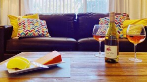 Un salon avec un canapé en cuir foncé, des coussins colorés, une table basse en bois sur laquelle se trouvent une bouteille de vin, des verres et une assiette de nourriture.
