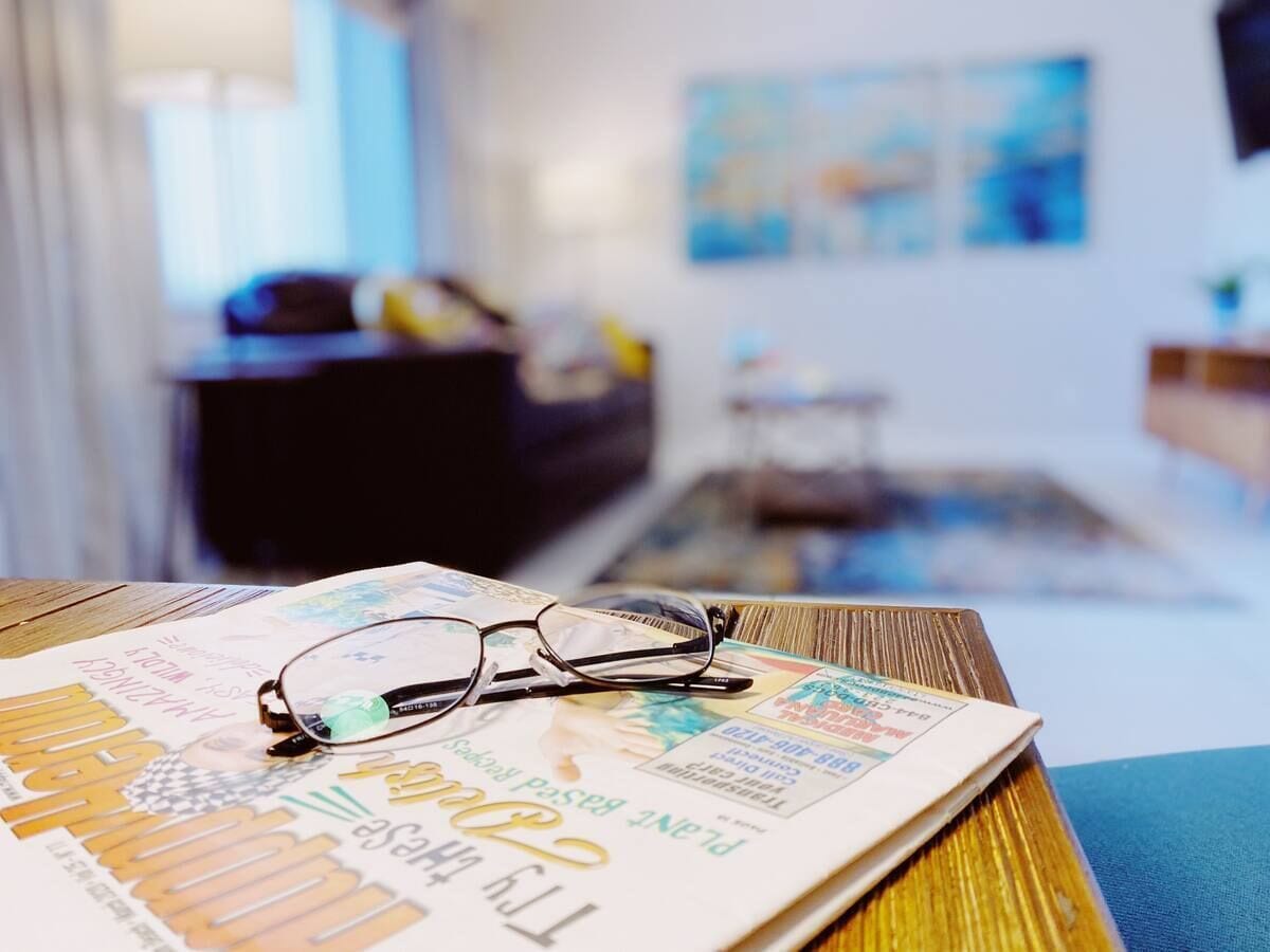 A pair of glasses on a magazine on a wooden table in a room with a blue wall and a window.