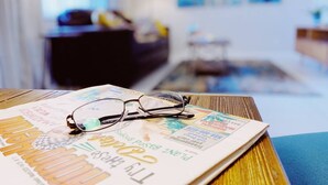 Une paire de lunettes posée sur un magazine, lui-même posé sur une table en bois, dans une pièce dont un mur est bleu et qui possède une fenêtre.