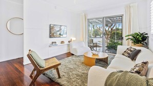 Ein modernes Wohnzimmer mit Sofa, Sessel und Couchtisch. Ein großes Fenster mit Vorhängen, eine Pflanze und Kunstwerke an der Wand.