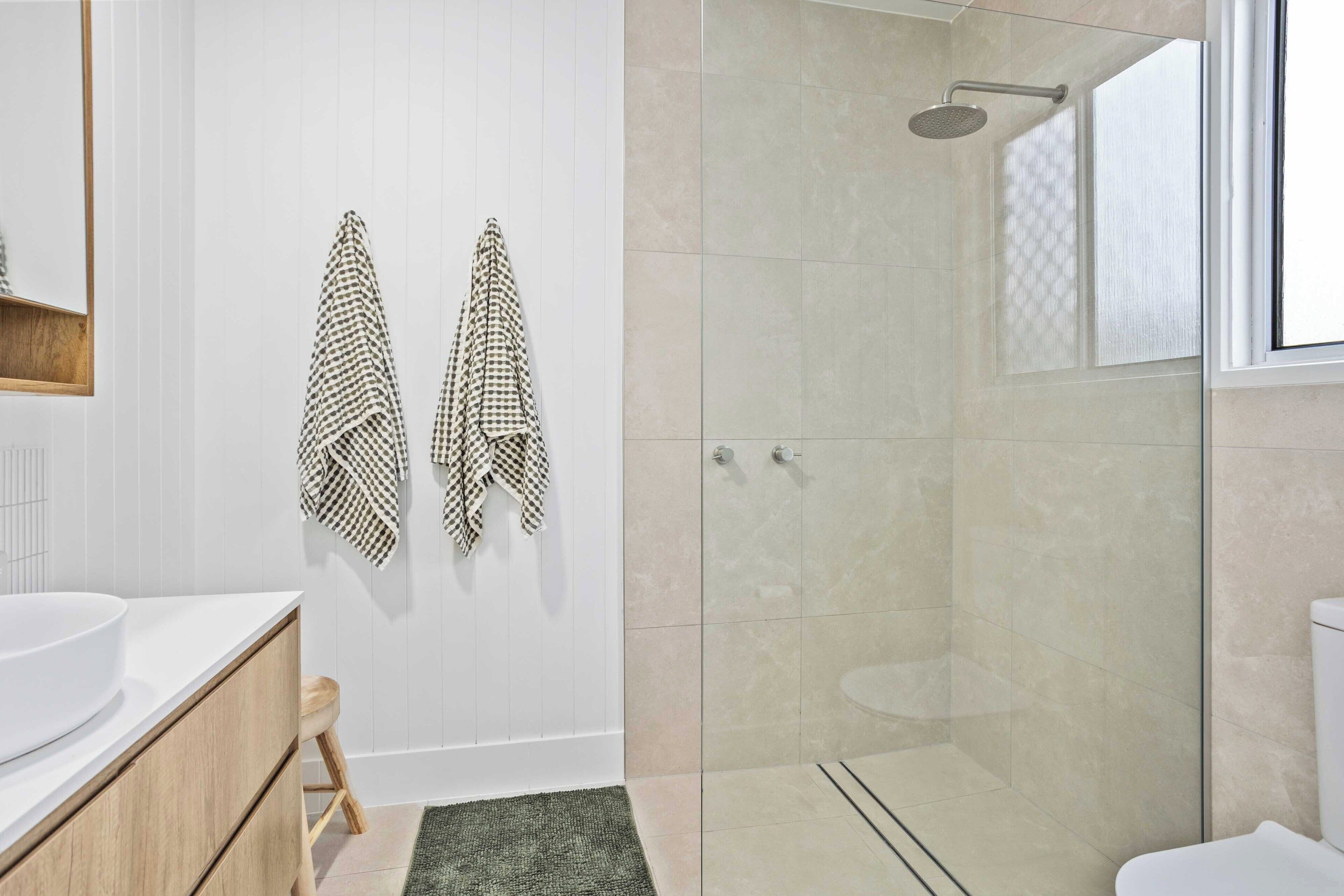A modern bathroom with a glass shower enclosure, a white sink, wooden cabinets, and two patterned towels hanging on the wall.