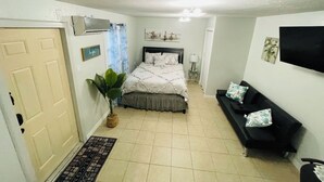 Un dormitorio con una cama, un sofá negro, una planta en maceta y un televisor en la pared.
