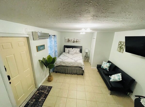 Un dormitorio con una cama, un sofá negro, una planta en maceta y un televisor en la pared.