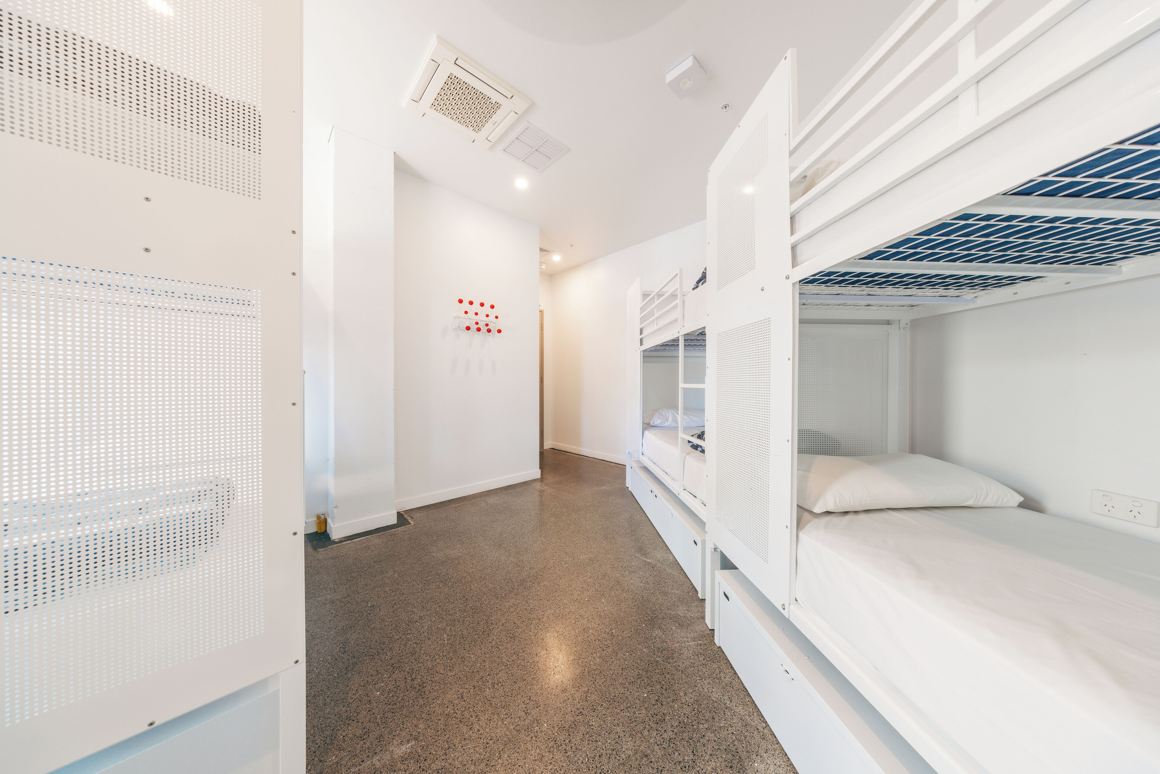 A dormitory room with bunk beds, a white wall with a red dot pattern, and a ceiling vent.