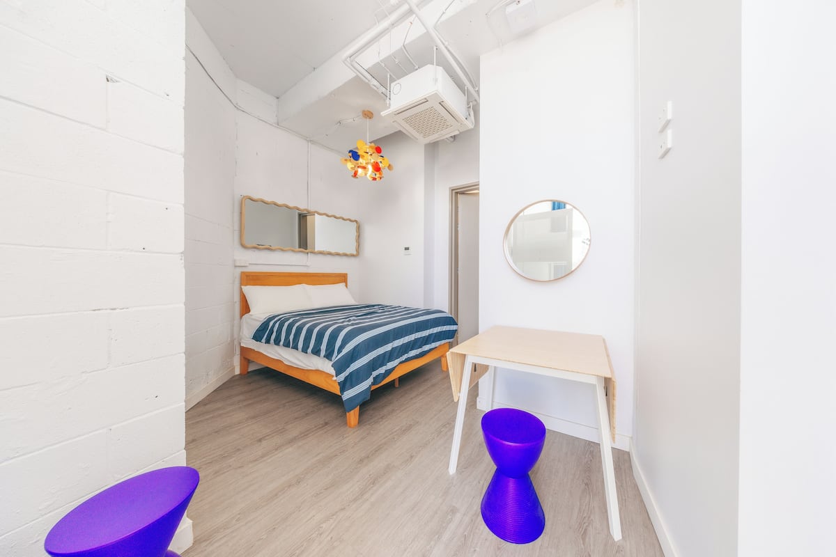 A bedroom with a bed, a desk, a chair, a mirror, and a hanging light fixture.