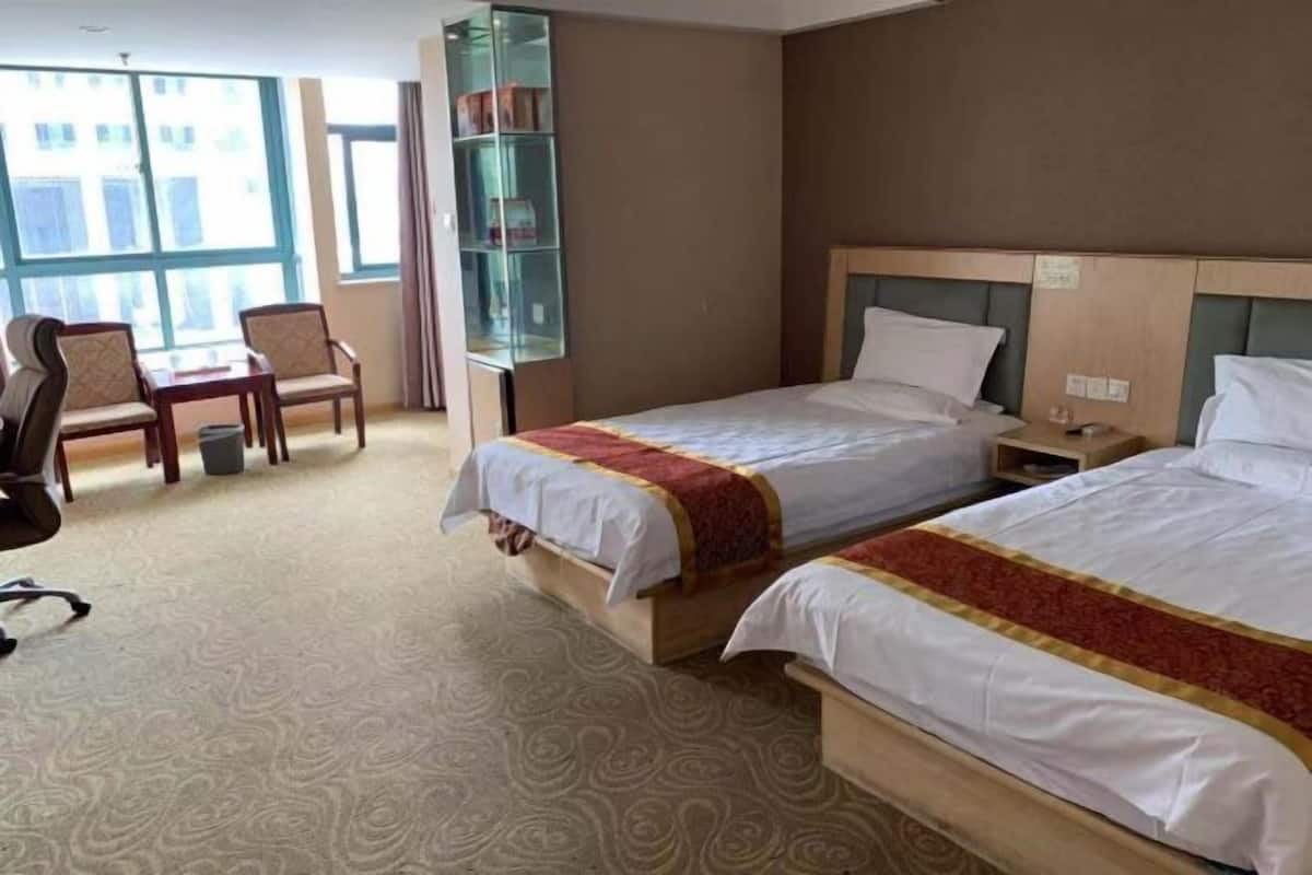 A hotel room with two beds, a desk, chairs, and a bookshelf.