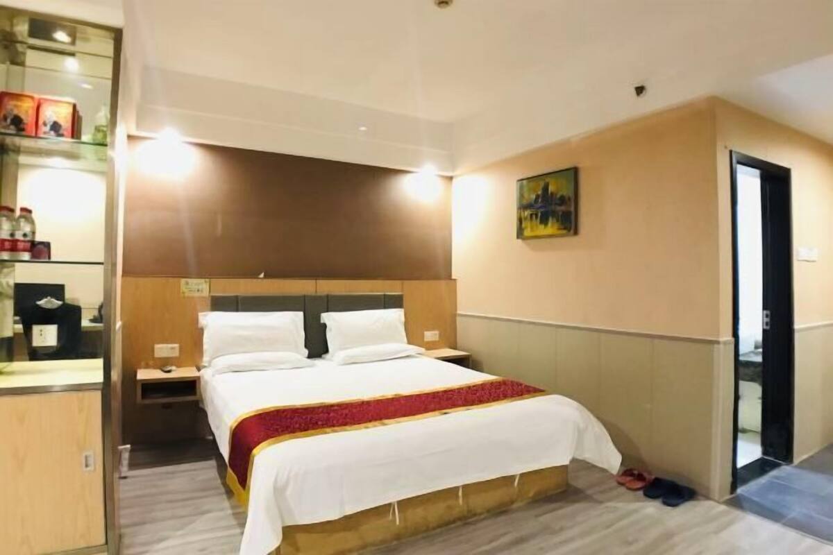 A hotel room with a bed, bedside tables, a nightstand, a painting on the wall, and a door leading to another room.