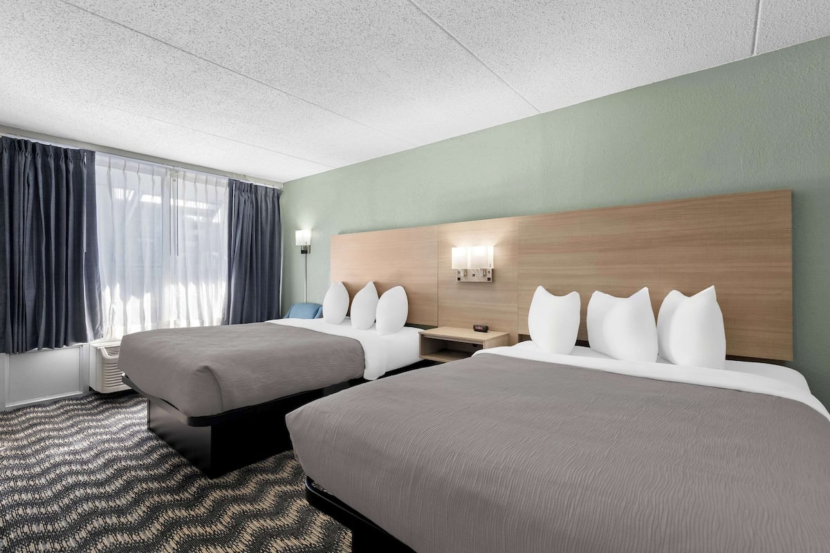 A hotel room with two beds, a wooden headboard, a nightstand, a lamp, and a window with curtains.