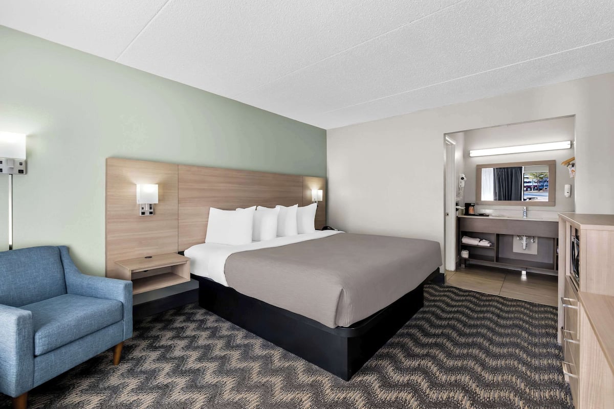 A hotel room with a bed, a desk, a chair, a vanity with a mirror, and a wall-mounted light fixture.